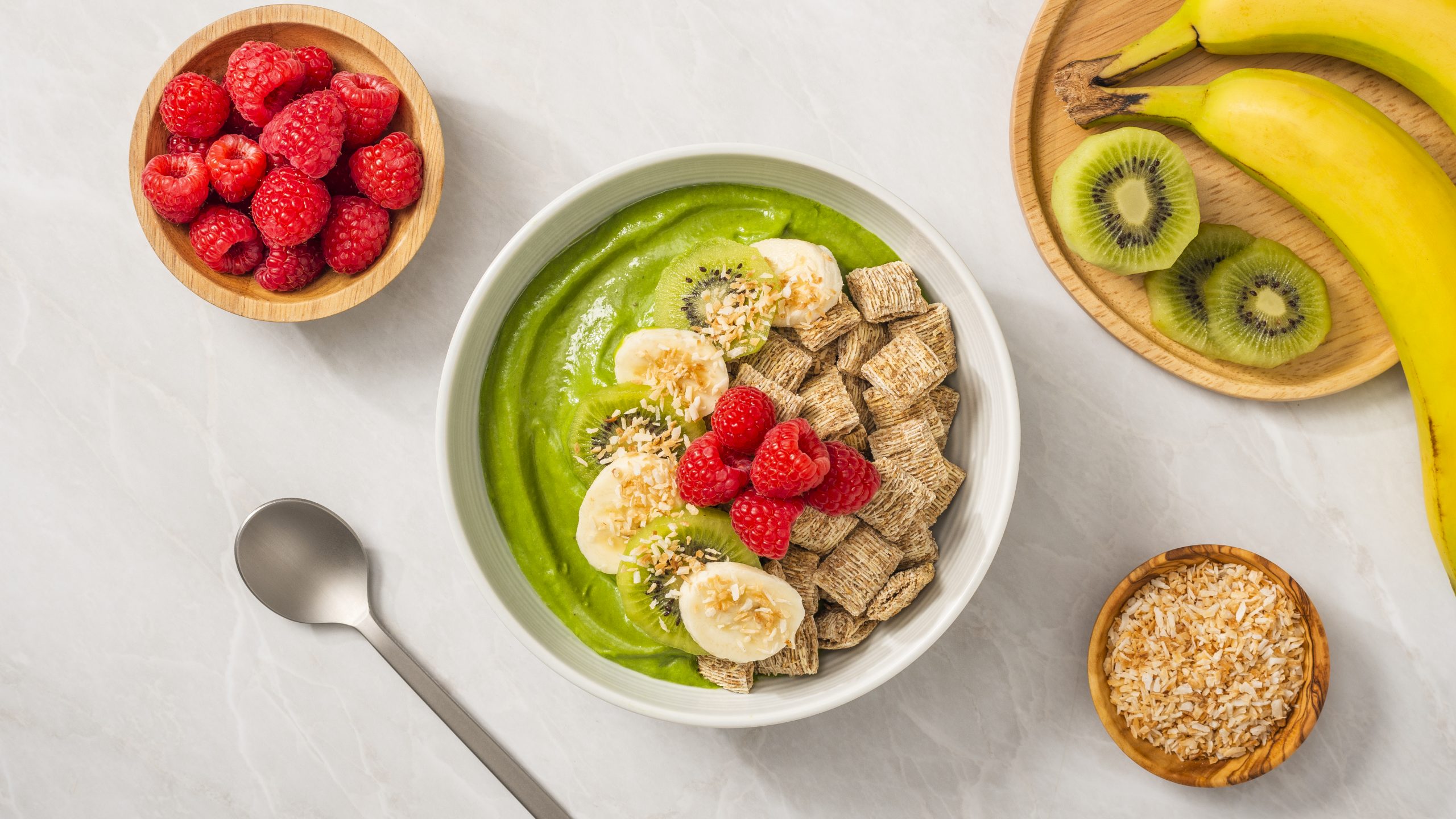 Green Goddess smoothie made with Shredded Wheat in a white bowl with bannana slices and raspberries on top. Sliced kiwi, banannas and raspberries in seperate bowls