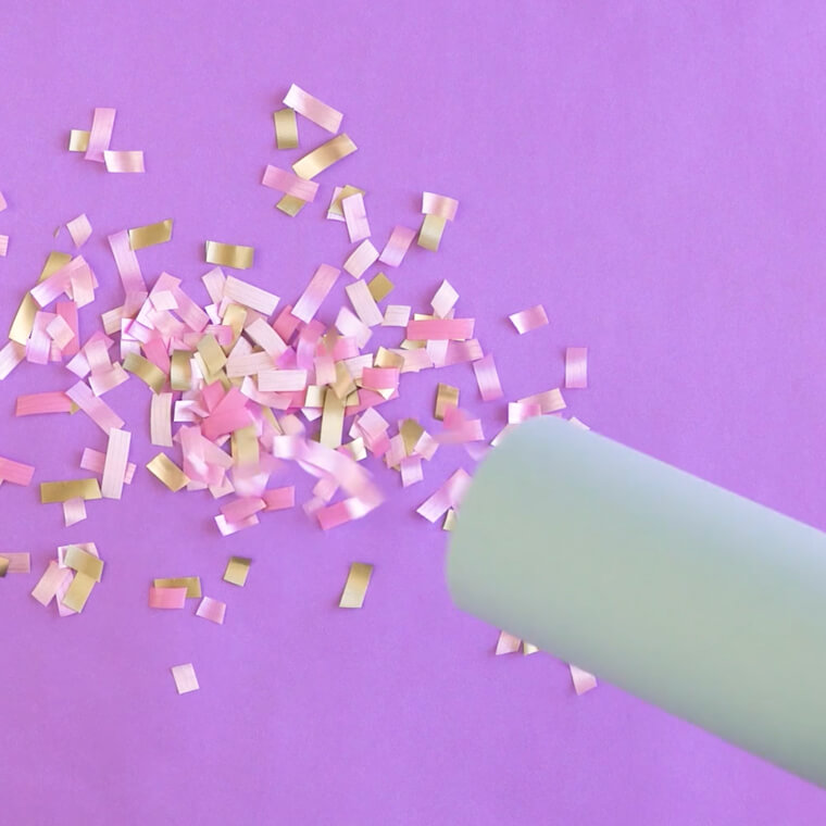 Pink, white and yellow confetti on a purple background being blown from a cardboard tube.