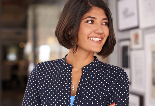 Woman in a blue and white dotted blouse with brown short hair smiling with her arms crossed. Let's Make Life Better slogan and an illustrated ribbon.